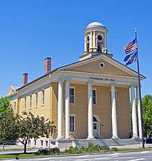 A pale-yellow two-story building, seen from the side, with a classically-styled colonnade on the front underneath a small domed cupola