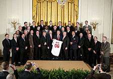 Three rows of men in various dark-colored suits; in the center, a gray-haired smiling man holds a white baseball jersey that reads "Bush" on the back in small red print with "06" in larger red print below it.