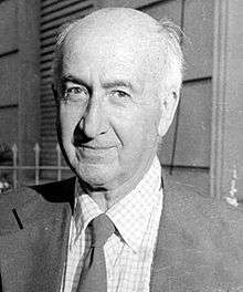 Black and white photo of a bald man looking straight wearing a suit and a tie