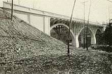 Black and white three-quarter view from just above water level of a triple span reinforced concrete, open spandrel arch bridge spanning a river.