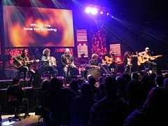 A group of seven musicians are sitting on a stage, performing with acoustic instruments