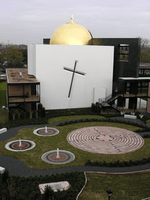 Concrete facade of the building is adorned only with a cross, etched at an angle, and a gold dome on top. Visible are external stairs, halls and lobbies. In the back is a standalone wall that bisects the dome, in which there is a window with bells.