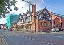 The front wing of the building seen from its corner.  It has two storeys; the lower storey is in brick with stone dressings and contains two wide arches in the entrance front; the upper storey is timber-framed and gabled; at both extremities there are tall chimney stacks with spiral decorative brickwork.  Behind the entrance wing are larger wings that contain the baths.