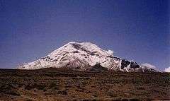 A snow-capped mountain lies in the distance against a cloudless blue sky. The land in the foreground is very barren.