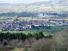 Multiple houses mostly with red roofs seen in a valley between the vegetation in the foreground and the hills beyond.