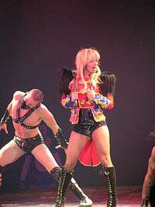 Image of a blond woman. She is standing with a red feathered jacket, carrying a whip around her neck and singing in a wireless microphone. Several people surround her, all wearing S&M outfits.