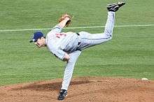 A man in a gray baseball uniform and navy-blue cap follows through after throwing a baseball. He is standing with his right leg planted on a dirt mound within a grass field and his left leg raised in the air behind him.