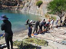 A group of about 12 divers on the shore of a flooded quarry preparing surface supplied diving equipment for diver training exercises. Several umbilicals are laid out for use in figure 8 coils.