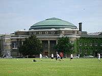 In the foreground is a grass field with seven male students playing soccer. A two-storey brick building is beyond the field, topped by an arched and nearly-triangular green-copper dome. A light grey-blue sky is visible in the top third of the image, with a helicopter in the distance on the right. Four automobiles are parked in front of the building along its length, and horizontally to it.