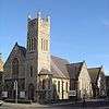 A pale stone church on a corner site, with a dominant square tower with spirelets. To the left of this is the entrance, below a four-light lancet window and three sexfoils.  To the right is the main body of the church, with two side chapels and lancet windows.