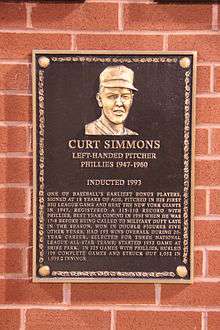 A dark bronze plaque with a man's face at the top and gold writing beneath, mounted on a brick wall