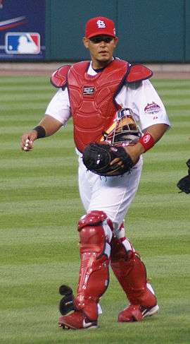 A baseball player walks in from the bullpen in his catcher's gear.