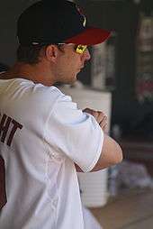 A profile view of a man in a navy blue baseball cap with a red brim; he is wearing mirrored sunglasses and a white baseball jersey