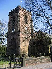 A square castellated tower viewed from one corner, with its front, incorporating a clock and an arched entrance, in shadow. The main church building, about half the height of the tower, is to the right and is connected to the tower.