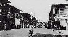 Man bicycling down street in old photo, with cars in background