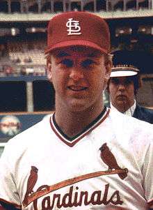 A man in a white pullover baseball jersey with "Cardinals" across the chest in red script, accompanied by a baseball bat and two male cardinal birds, and a red baseball cap with a white interlocking "StL"