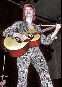 A color photograph of David Bowie with an acoustic guitar