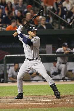 A man in a gray baseball jersey and a navy blue batting helmet stands in a batters box, preparing to swing at a pitch.