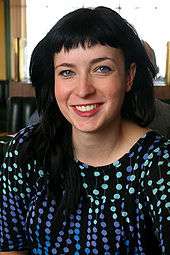 Profile of a smiling woman with black hair who is wearing a black blouse with blue and green dots.