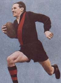 Wearing a black long sleeved jersey with a red stripe and black shorts, a young male athlete strides with a football wedged in one arm.