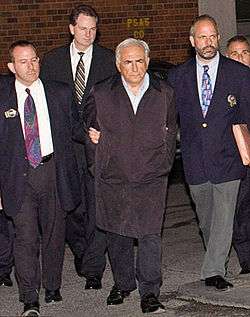 A white-haired man in a black overcoat and dress shirt with his hands behind his back at the center of a small group of men walking toward the camera. The two men on either side are wearing jackets with gold badges clipped to the lapels and ties. They are holding the arms of the man in the center. A fourth man, also in a jacket and tie, is visible in the rear.