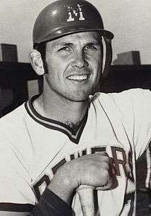 A black and white photograph of a man wearing a white baseball jersey with "Brewers" written across the chest and a batting helmet with an "M" printed on it.