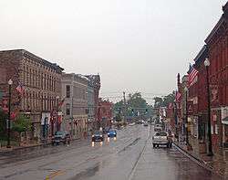 A wide, wet street with tall commercial buildings on either side. Two approaching vehicles have their headlights on, and a traffic light behind them is green. In the rear of the image the street narrows to go over a small bridge.