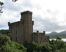 A grey castle with tall square towers stands amongst trees in full leaf.