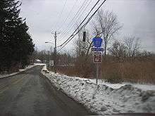 A rural, narrow two-lane highway passes through an area composed of trees, fields, and small brush.