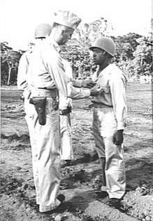 Tall man wearing garrison hat and a pistol in a holster pins a medal on short man wearing steel helmet.