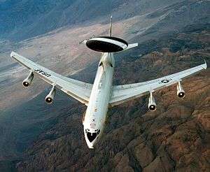 Aerial view port view of white jet aircraft in-flight. It has a large disc-like black radar lying horizontally above two convergent struts.