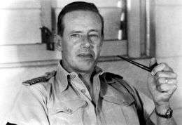 Informal portrait of man with short dark hair in light-coloured open-necked military shirt, holding a pipe