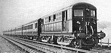 A black and white image of an electric locomotive hauling at least 6 coaches, shown with the electric locomotive on the right. There is an track in the foreground that is electricifed with the fourth rail system, the locomotive is shown with two pickup shoes.