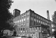 A five-storey cuboid factory composed of brick and windows. The view is from the ground at one corner of the factory. To the right, appearing from behind the factory, is a tall brick chimney. To the left are trees appearing black, and at their fringe, a square brick tower connected to the factory.