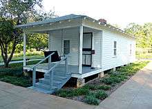 Present-day photograph of a whitewashed house, about 15 feet wide. Four bannistered steps in the foreground lead up to a roofed porch that holds a swing wide enough for two. The front of the house has a door and a single paned window. The visible side of the house, about 30 feet long, has two paned windows.