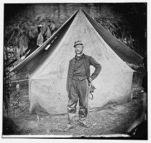 A white man with a mustache and long sideburns standing before a military tent. He is wearing a military jacket over a vest and tie, a sword at his belt, and a kepi on his head.