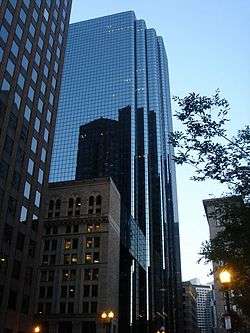 Ground-level view of a 40-story skyscraper with an all-glass facade and several setbacks. In the facade is a reflection of a dark brown building with lighter windows, and near the bottom of the skyscraper is a small white building with dark windows