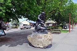 A cast sculpture mounted on a boulder. The sculpture depicts a long haired and bearded man, dressed in a shirt and trousers and carrying a staff. By his side is a sheep dog. Both the man and the dog are facing right. A brass plaque is on the boulder reading "James MacKenzie & dog