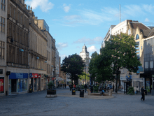 Fargate shopping precinct, Sheffield. Once a busy road, it has been pedestrianised for several decades and is Sheffield's main City Centre shopping area, home to many well-known companies. The image shows classical architecture on both sides with one plan spaces in the centre, dotted with trees and the buildings on the High Street are visible beyond the trees.