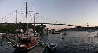 A docked sailboat floats in front of a suspension bridge, at twilight.