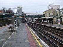 A view along a station platform. The main station building is to the right across a pair of tracks. A footbridge spans between the platforms.