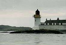 A small white lighthouse with a black canopy sits on a dark rock. There is a high white wall and a row of black and white houses to the right and dark hills at left in the distance.