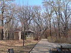 Boardwalk running through woods in fall; wooden picnic table and wood display structures beside trail