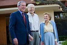 A man in a suit is standing next to an older man and woman in casual attire. The trio stands in front of a yellow house.