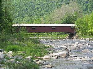 A red, wooden covered bridge over a rocky stream with a forest-covered mountain in the background