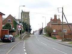 A narrow road up a slight incline between red brick buildings of a variety of styles and ages. The pavement on the left is raised above the roadway. Left on the skyline is the square greenish church tower.