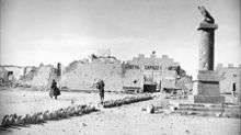 Two allied soldiers, in greatcoats, walk towards a bullet ridden and partially destroyed wall topped with crenelations. A bullet ridden column stands in the foreground.