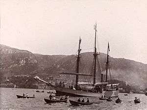 A three-masted ship, under steam power, moves across a stretch of water attended by several rowing boats. In the background is a line of hills, with buildings faintly visible at the water's edge.