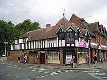 A two-storey building seen from the corner; the lower storey is brick and the upper storeys timber-framed.  The left side extends further than the right and contains the public conveniences, with two doorways on the ground floor and a long dormer above.  On the corner is a doorway with windows on each side; above are three windows extending round the corner, each with a gable.  Behind these is a tiled spire.