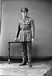 A full length portrait of a man in military uniform wearing medals. His hand is resting atop a small table.